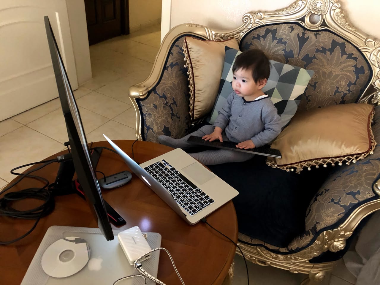 Zuhair's toddler son perched in an armchair, holding a laptop in front of a desktop monitor at a makeshift home workstation